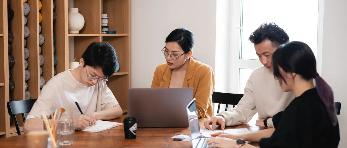 Vier collega's met laptops aan een houten tafel in een klein kantoor